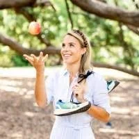 A woman in a white outfit and tiara playing with a pink ball outdoors among trees.