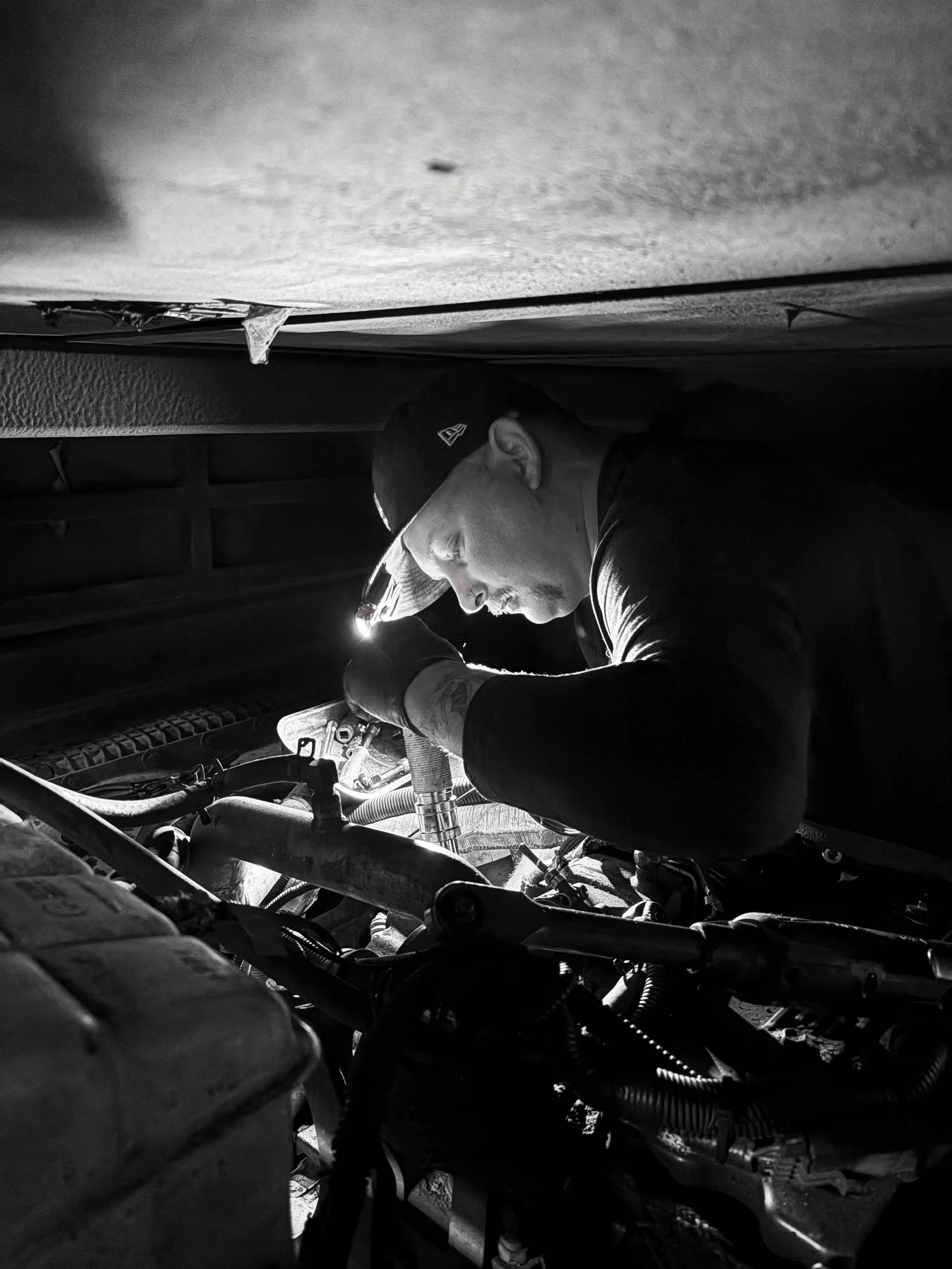 A man wearing a cap and gloves working on a vehicle engine in a dimly lit space, possibly a garage or workshop.