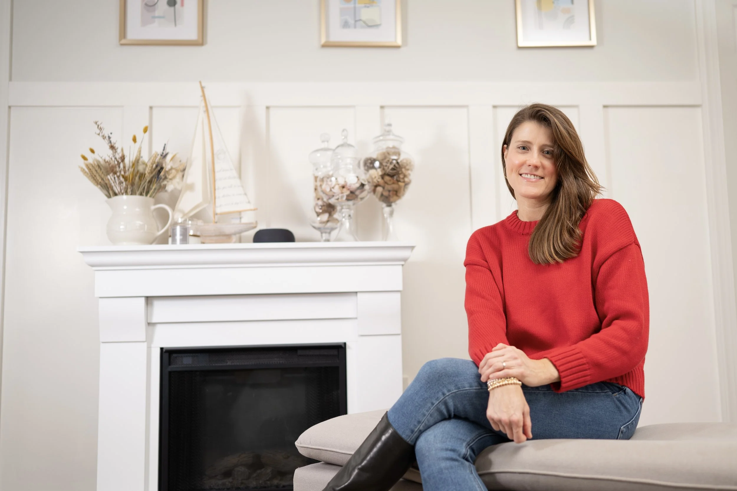 Woman sitting on a beige couch near a white fireplace, smiling, wearing a red sweater and blue jeans, in a living room with framed artwork and decorative items.