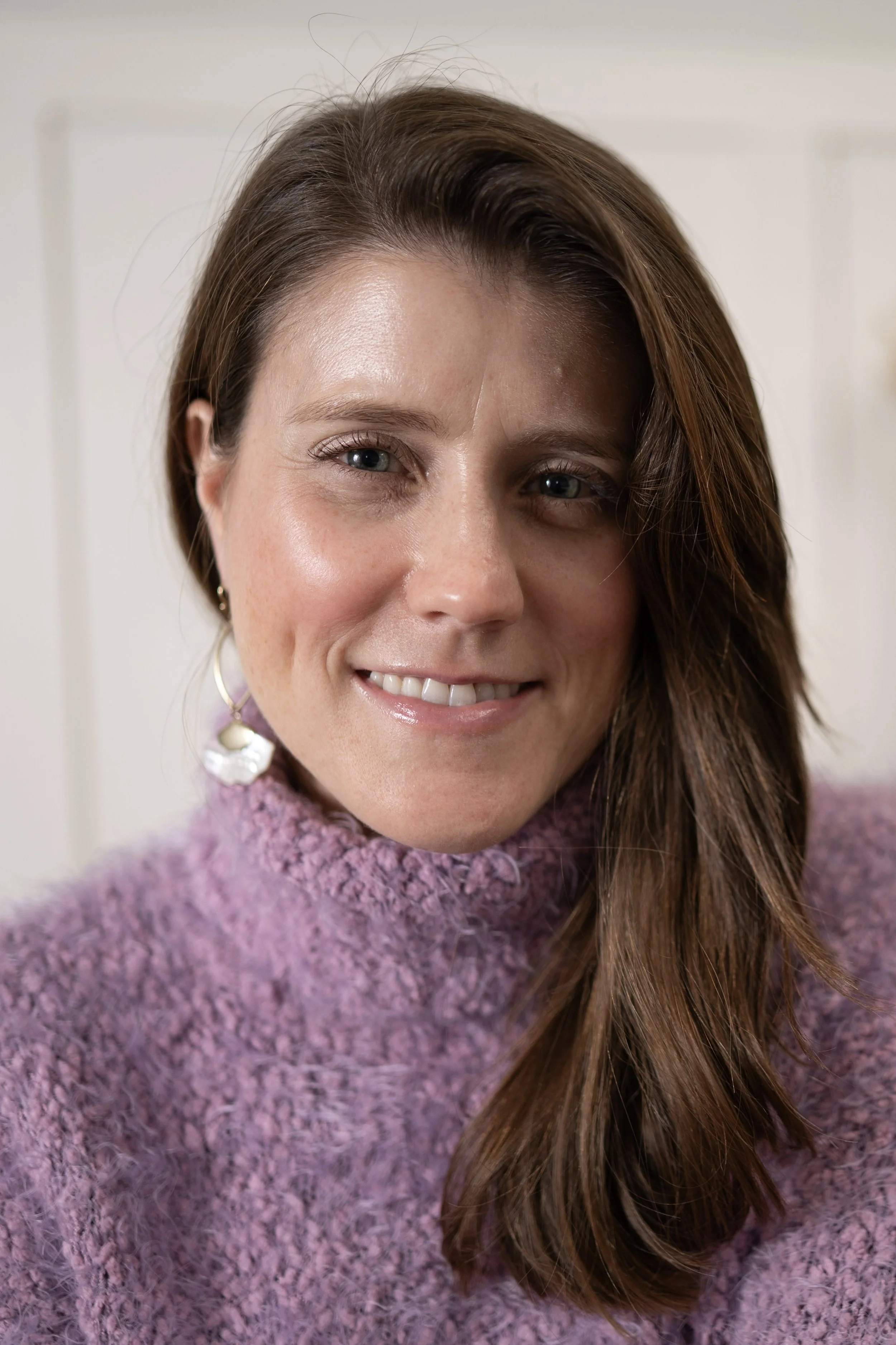 Close-up of a woman with shoulder-length brown hair, blue eyes, and wearing a purple fuzzy sweater, smiling at the camera.