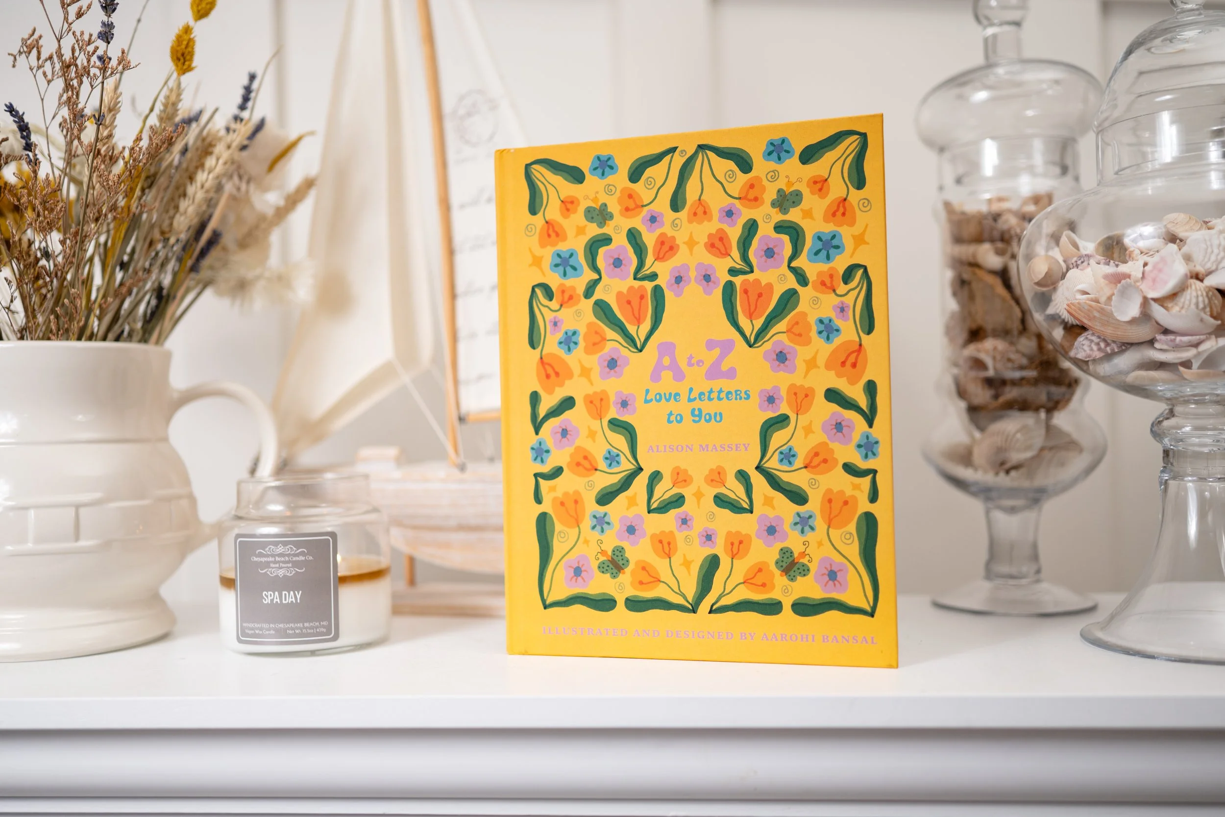 Book titled 'A - Z Love Letters to You' by Alison Massey standing on a white surface, surrounded by decorative jars filled with seashells and dried flowers.