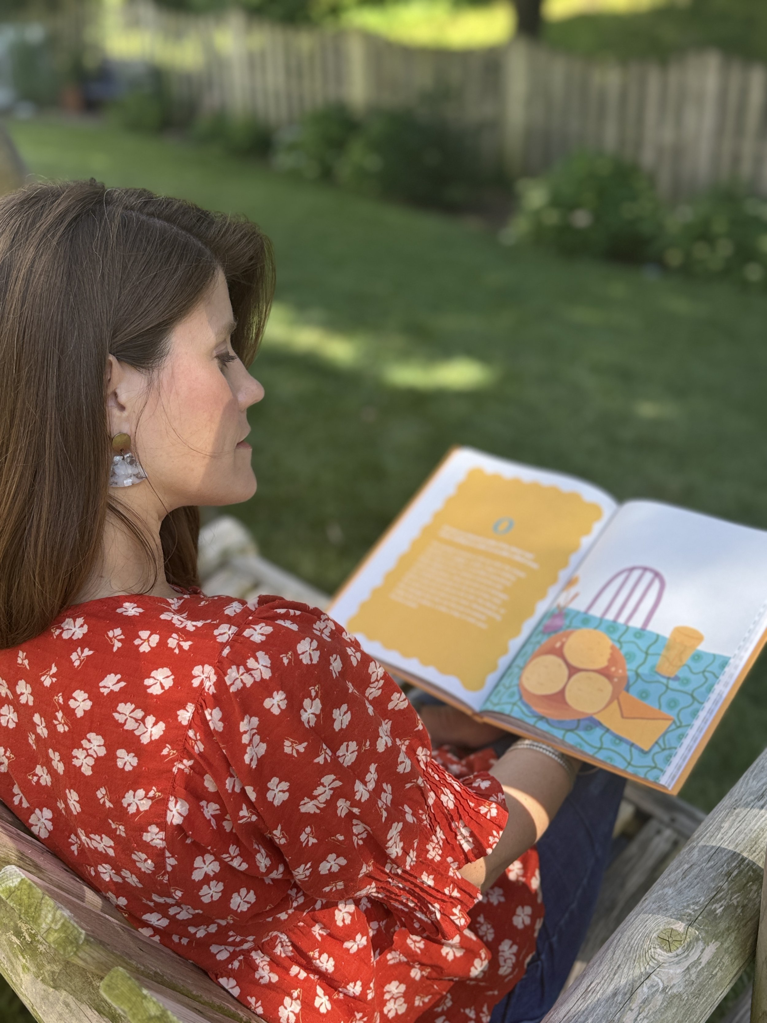 A woman with brown hair and earrings, wearing a red dress with white flowers, is sitting outside on a wooden chair, reading a colorful book with a yellow and blue illustration and a story on the facing page, in a garden with green grass and plants.