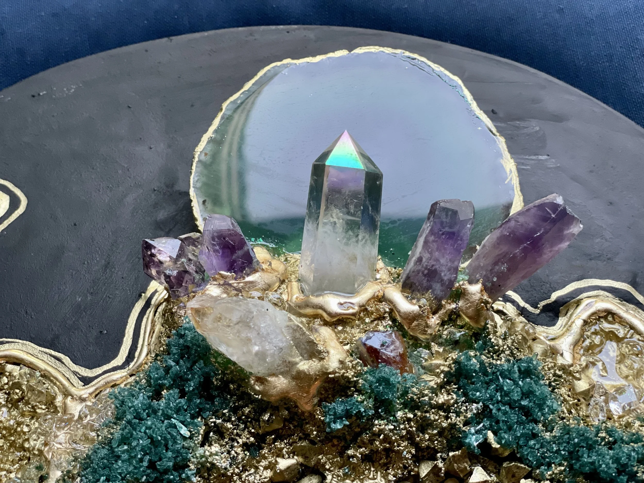 Close-up of a decorative piece featuring an open geode with crystals, a vertical quartz crystal, and various purple and clear crystals, set against a dark background with gold accents and textured teal elements.