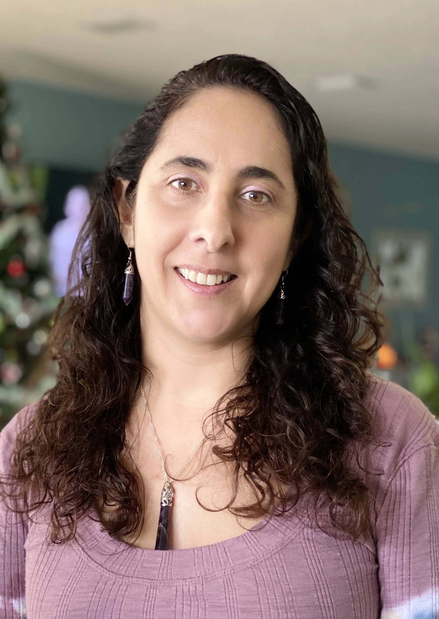 A woman with long, curly dark hair smiling, wearing purple earrings and a matching purple top, with a decorated Christmas tree in the background.
