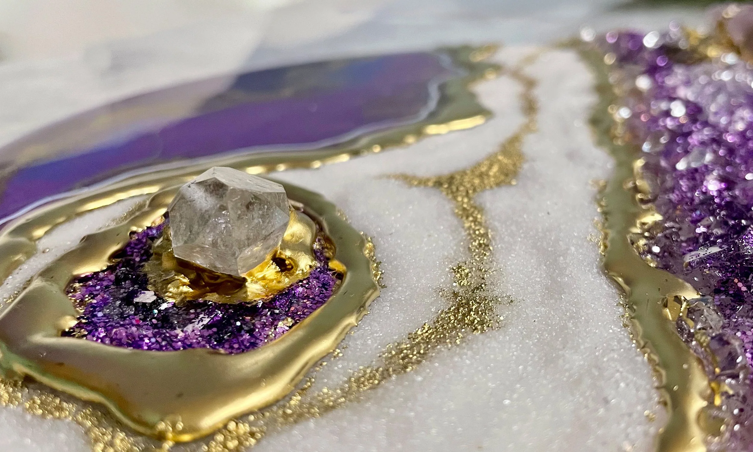Close-up of an abstract art piece featuring a clear gemstone, purple glitter, gold accents, and textured materials on a marble-like surface.