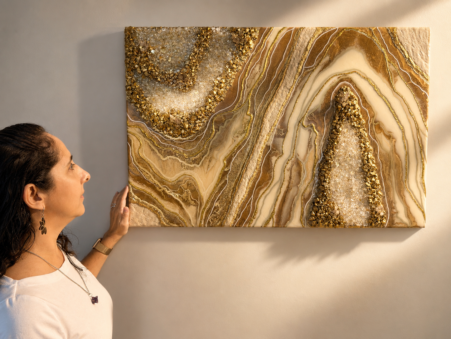 A woman with dark hair, wearing earrings and a watch, looks at a mixed media abstract art piece on a wall. The art features swirling layers of beige, brown, and cream tones with embedded textured gold and clear crystal-like elements.