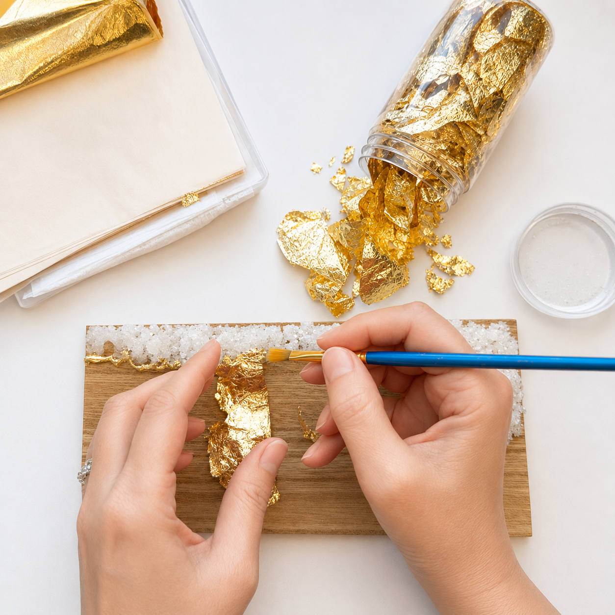 Person using a small brush to apply gold leaf onto a piece of wood. Gold leaf is stored in a clear plastic container, with some pieces on a salt block and others scattered on the table. There are additional sheets of gold leaf and paper nearby.