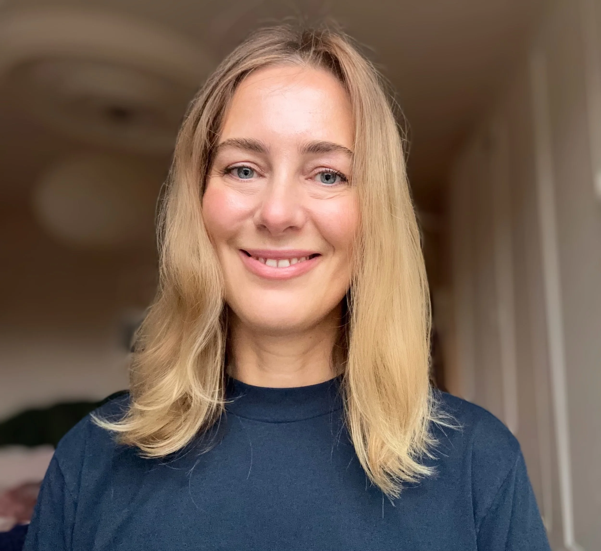 A woman with blonde hair, blue eyes, and light skin smiling and wearing a dark blue shirt, indoors with a blurred background.