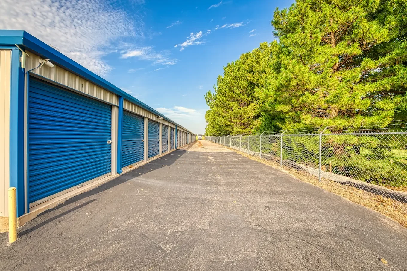A row of blue storage units with metal roll-up doors on the left, a chain-link fence on the right, and green trees behind the fence, under a partly cloudy sky.