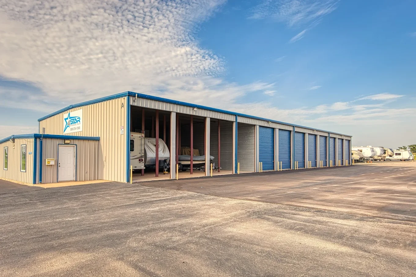 Large storage building with blue and beige exterior, partially open for parking boats, with a row of RVs parked in the background under a partly cloudy sky.