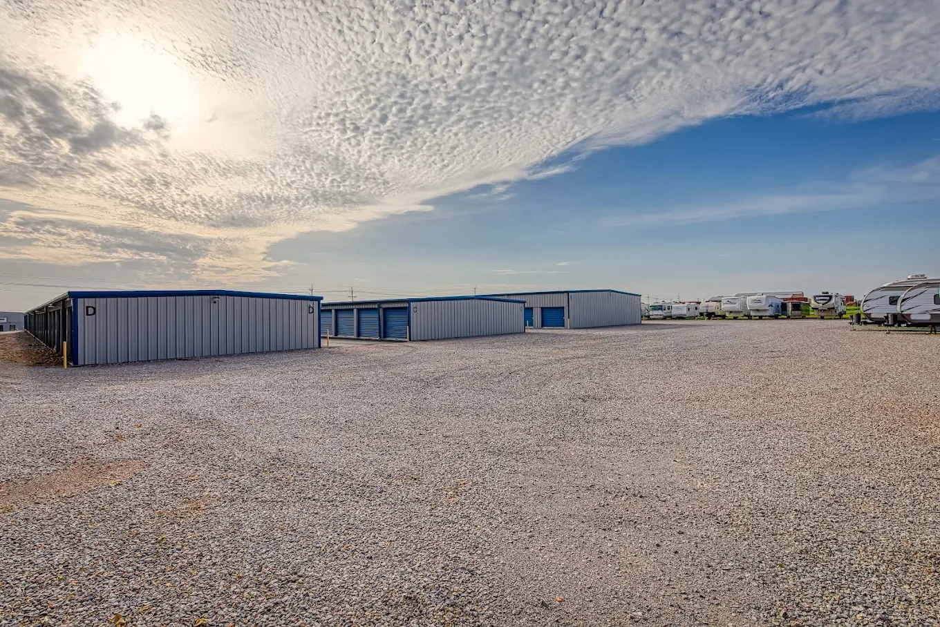 A wide outdoor storage lot with metal storage units on the left and RV trailers on the right, under a sky with partly cloudy and partly clear conditions.