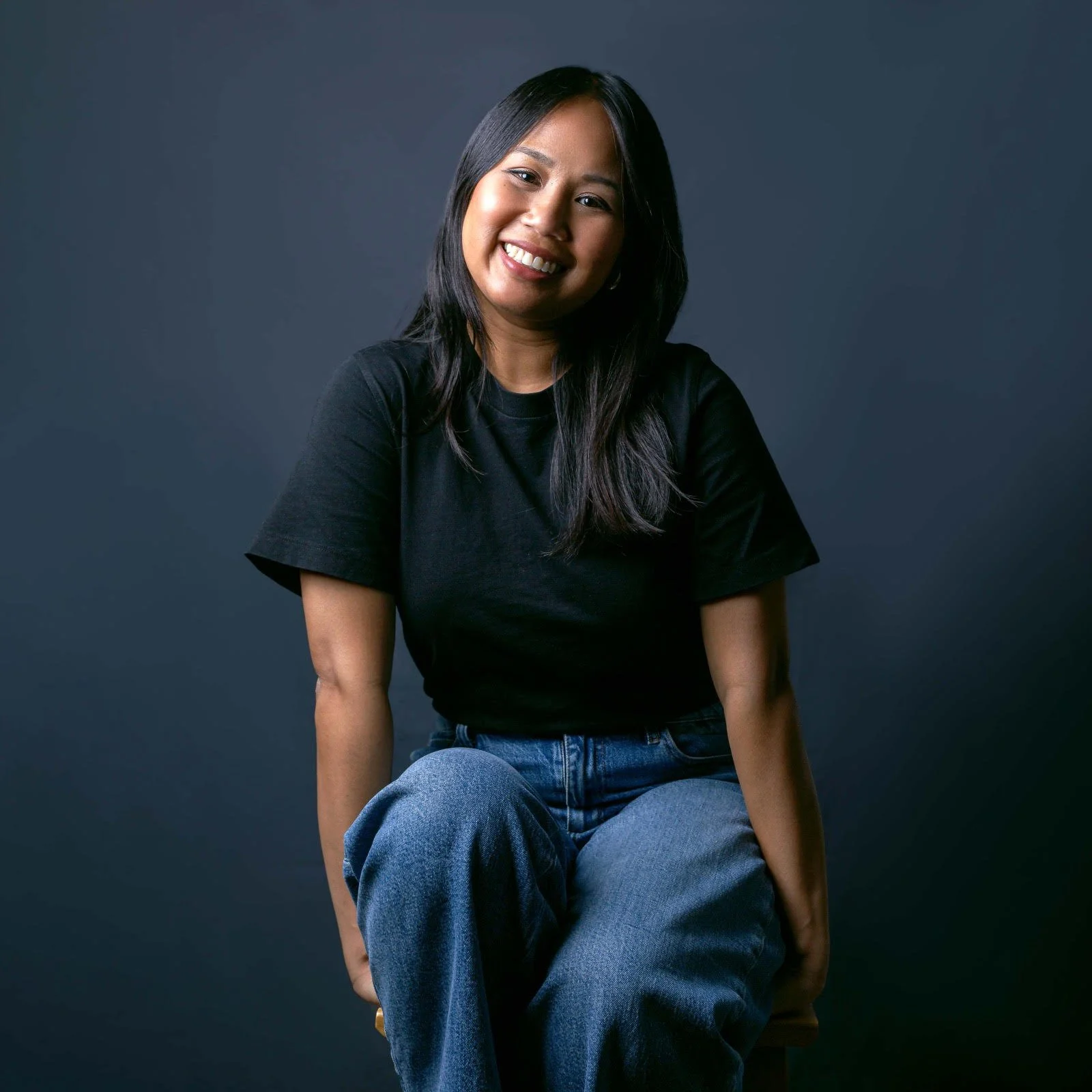A woman with long dark hair, smiling, wearing a black t-shirt and blue jeans, sitting against a dark gray background.