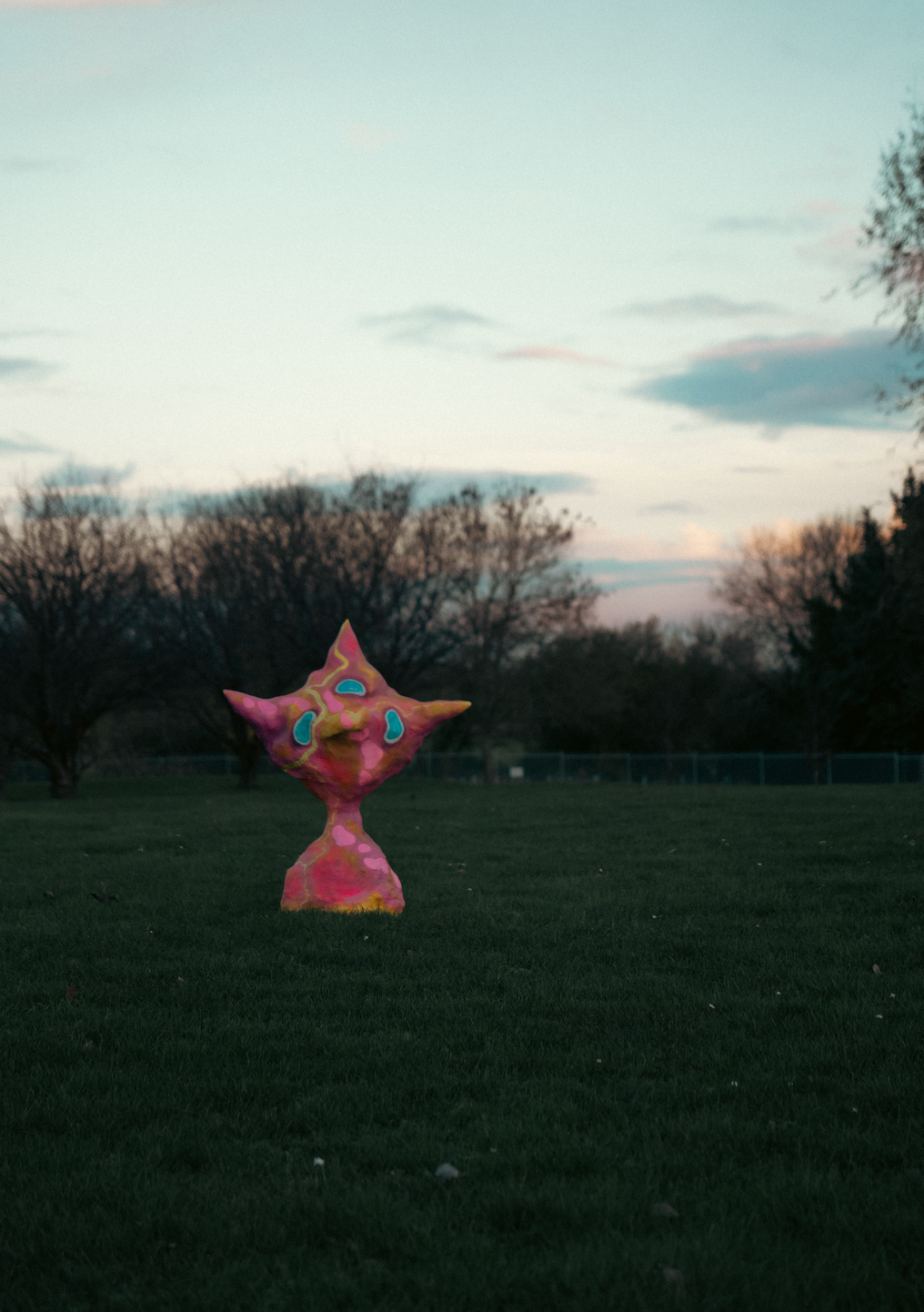 A colorful, abstract sculpture shaped like a cat standing on grass in an outdoor park during dusk, with trees and a cloudy sky in the background.