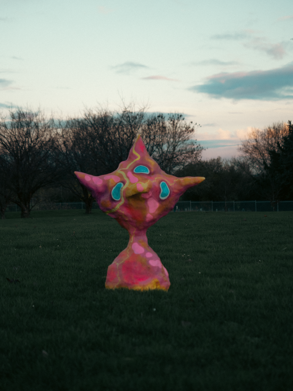 Colorful, abstract sculpture resembling a whimsical creature with multiple extensions, situated on a grassy field with trees and a cloudy sky in the background.
