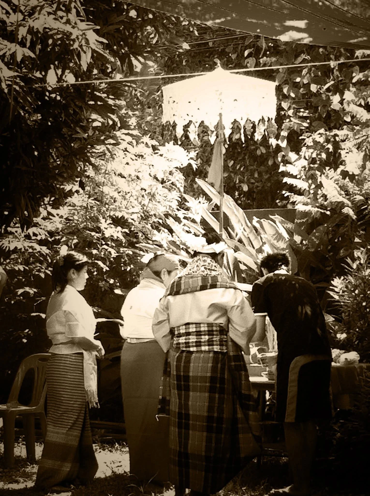 A group of people participating in a traditional outdoor ritual or ceremony surrounded by lush dense greenery and plants, with a thatched roof structure in the background.