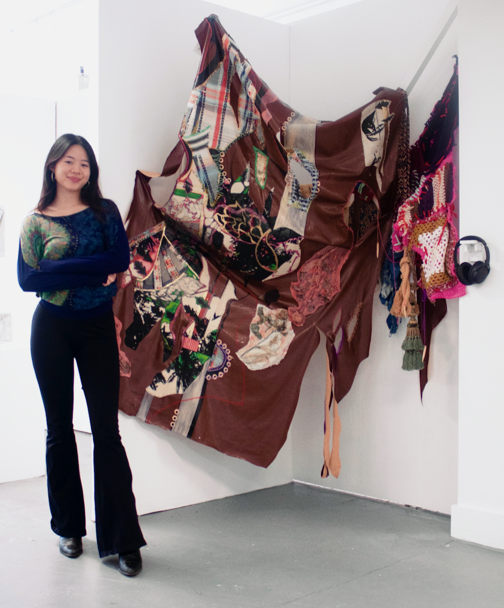 A woman standing next to a large, mixed-media textile art installation on a white gallery wall. The textile piece includes various fabric patches, embroidery, and accessories.