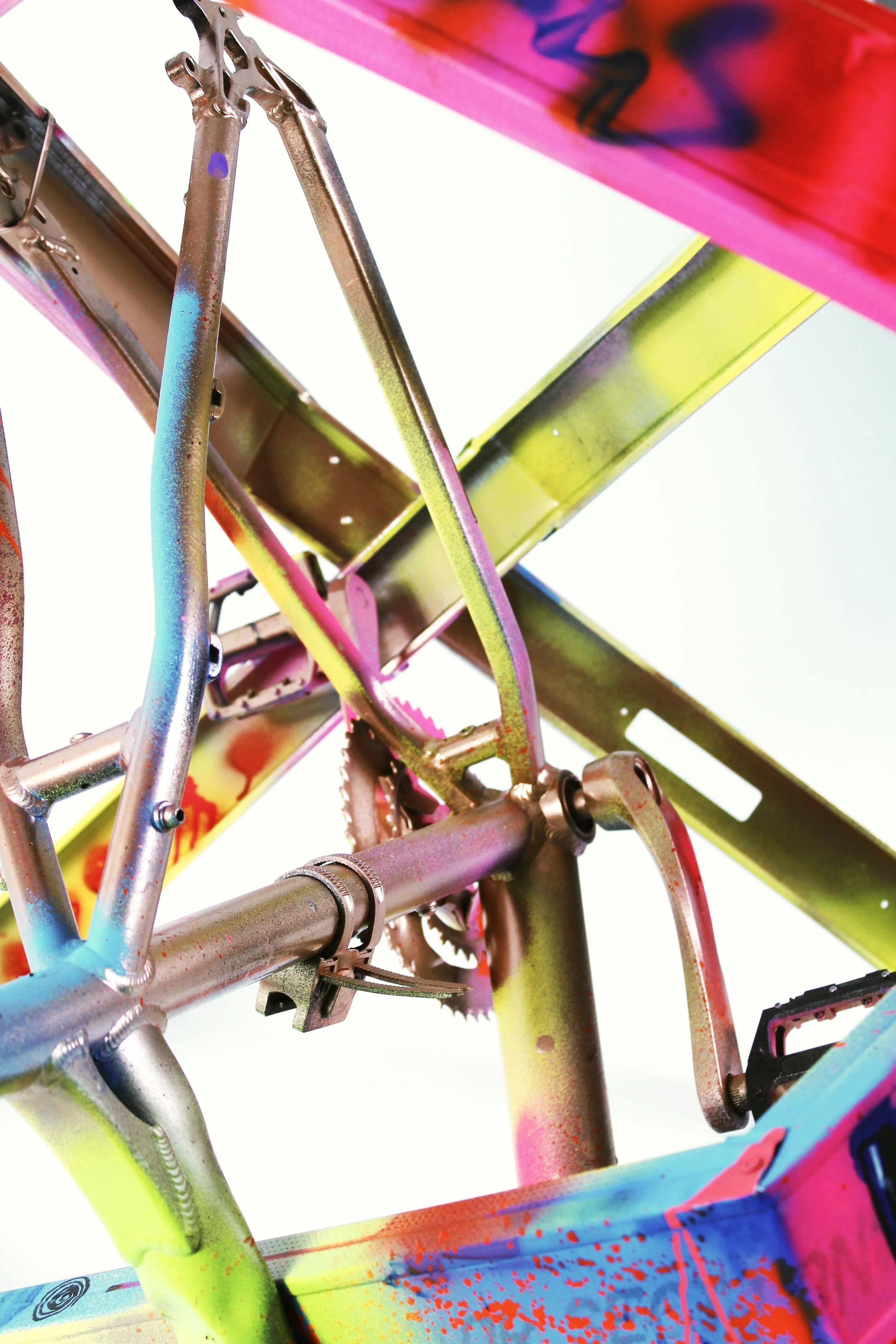 Close-up of a colorful, graffiti-painted bicycle frame with visible gears and pedals, shot from below against a plain background.