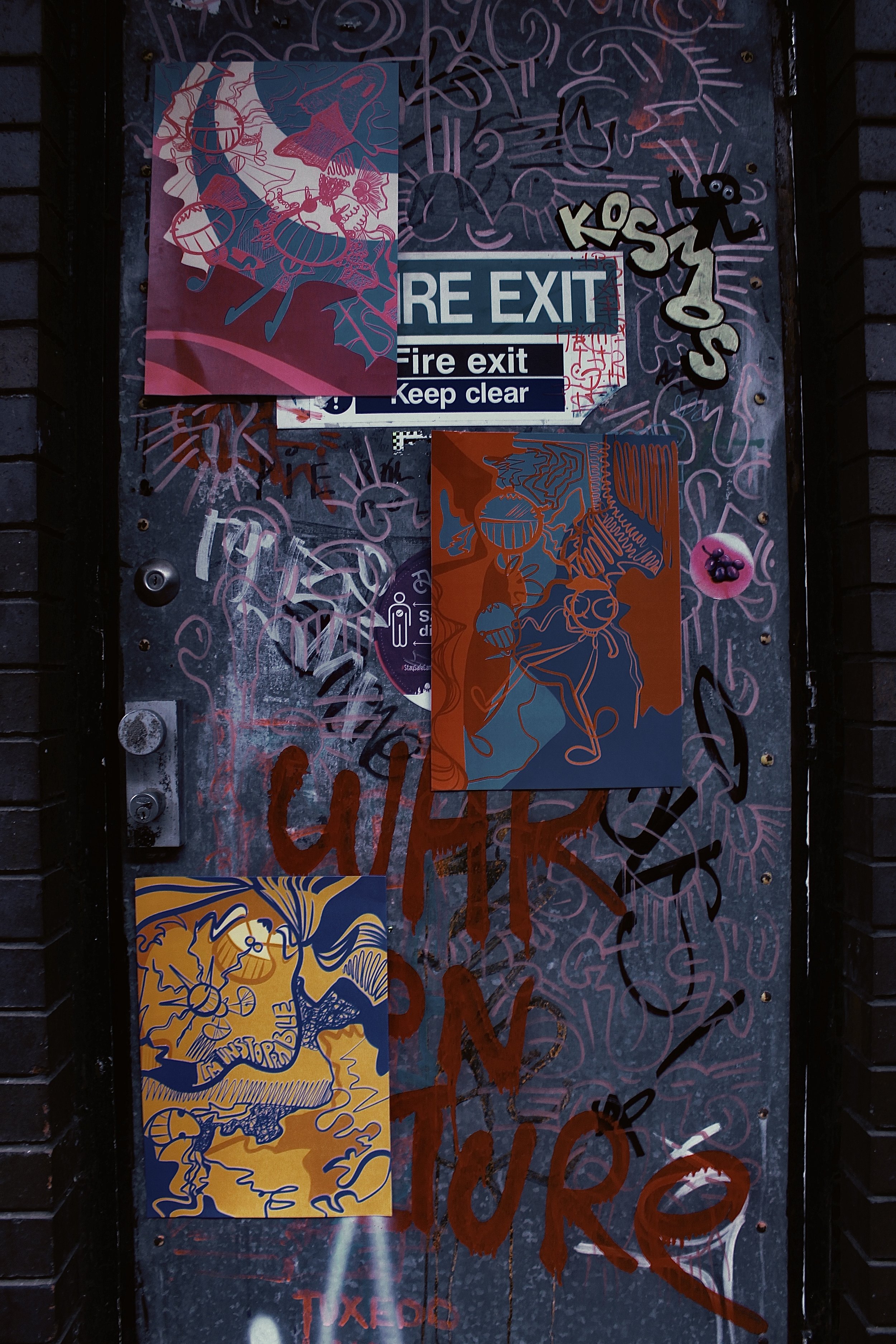 Graffiti-covered door with multiple posters and stickers, including a blue exit sign, colorful drawings, and red spray paint words. The door is surrounded by brick walls.