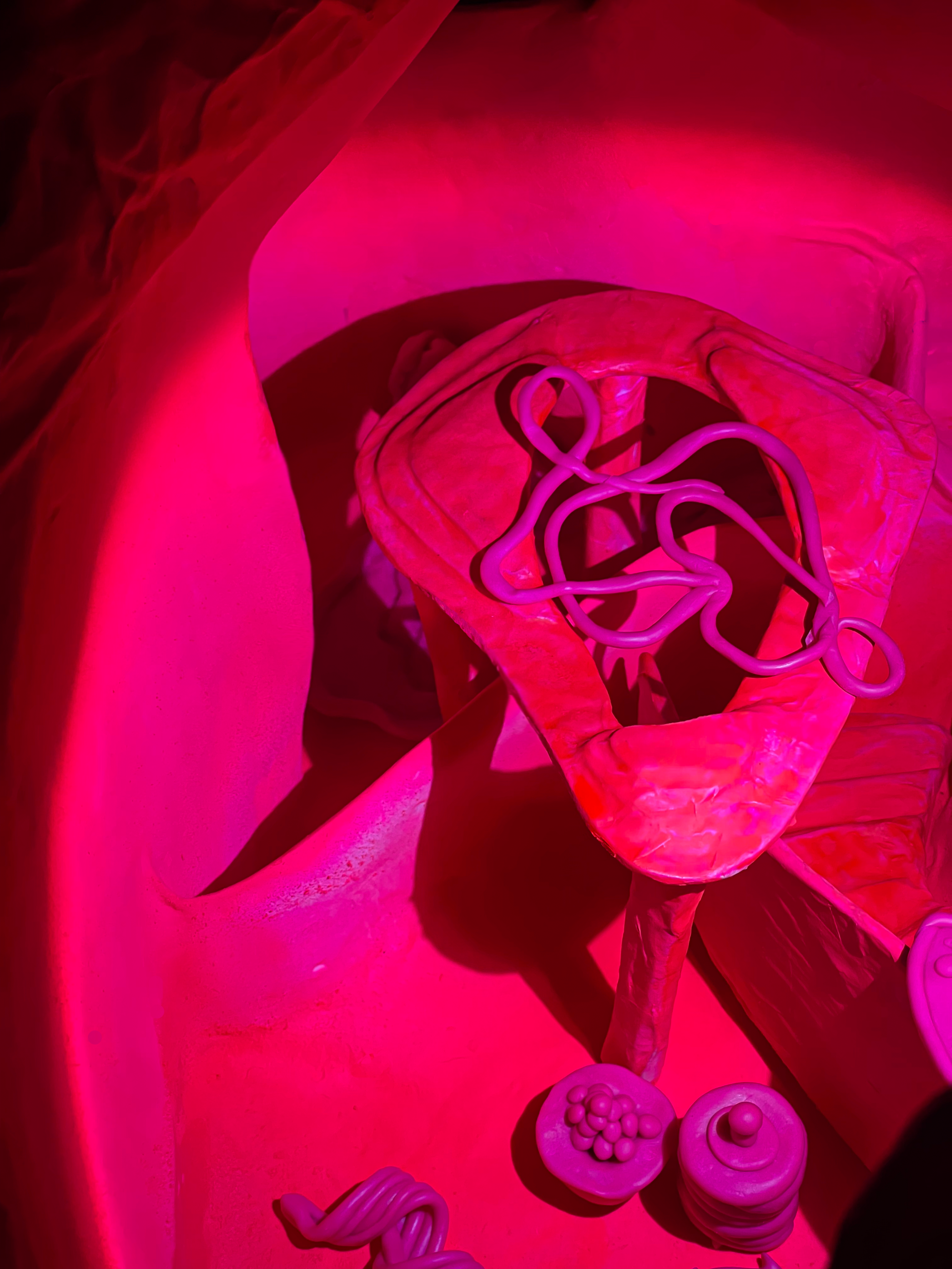 Pink-lit art installation with abstract shapes, a cloud-like structure, and small objects resembling food or toys.