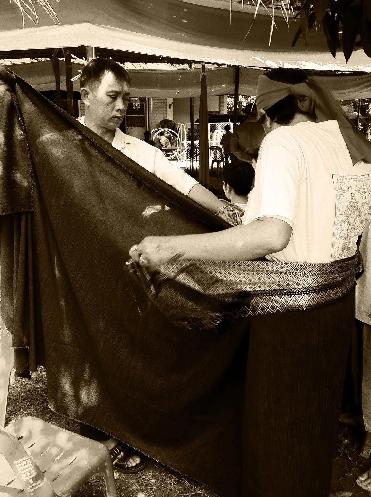 A man with light skin and short hair, and another man wearing a headscarf, are engaged in a transaction at an outdoor market stall. The stall has a fabric cover and various items for sale.