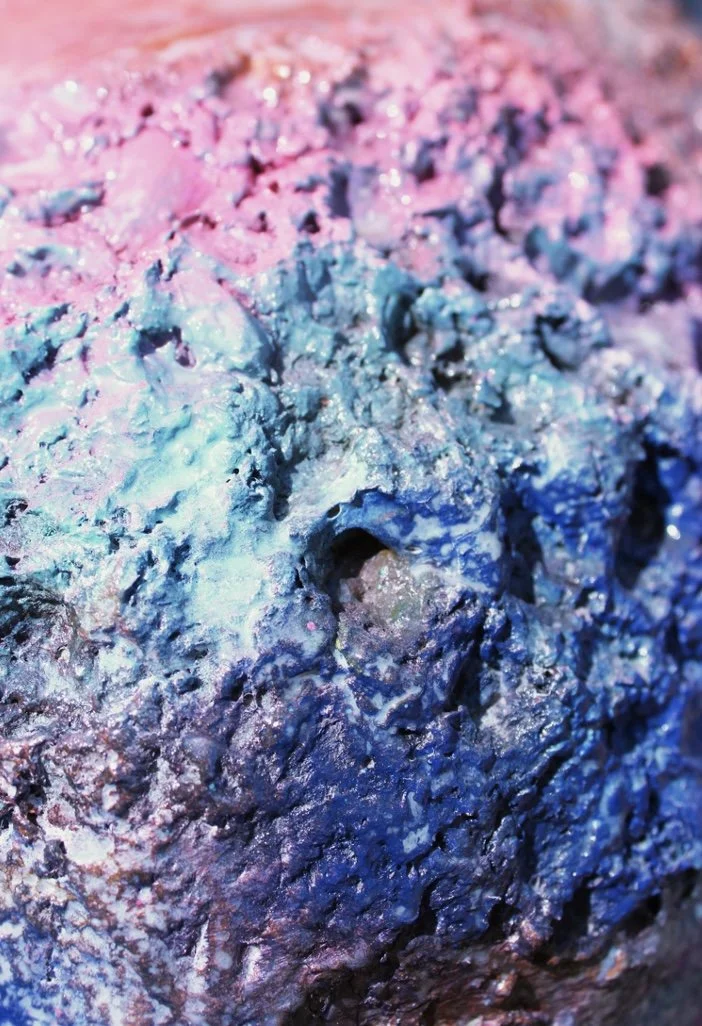 Close-up of a textured, multicolored surface with hues of pink, blue, and purple, resembling a rough, rocky surface with metallic and iridescent highlights.