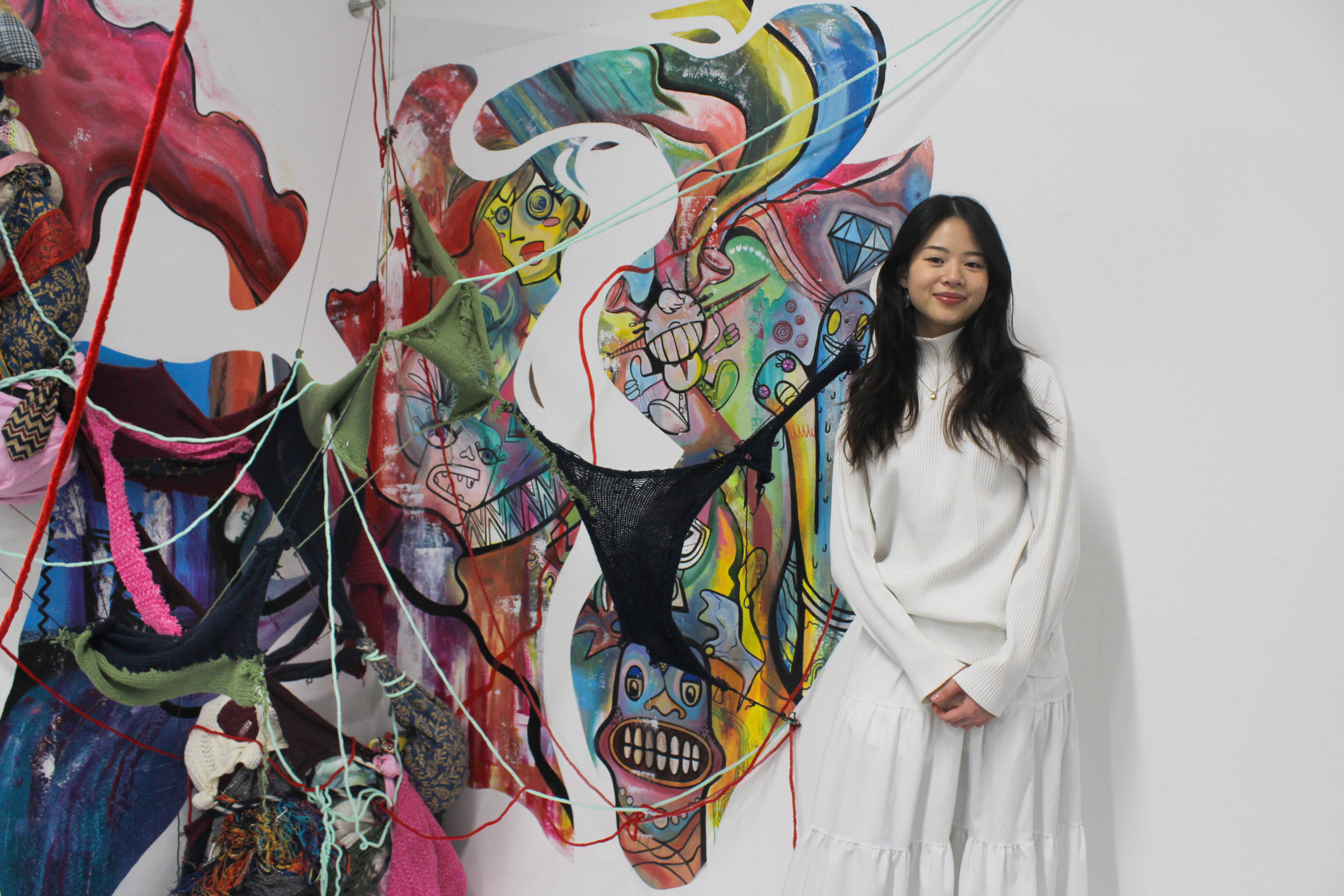 A young woman with long dark hair smiling in front of a colorful abstract mural. The mural includes various cartoonish faces, patterns, and shapes, with some knitted or cloth elements attached to it.