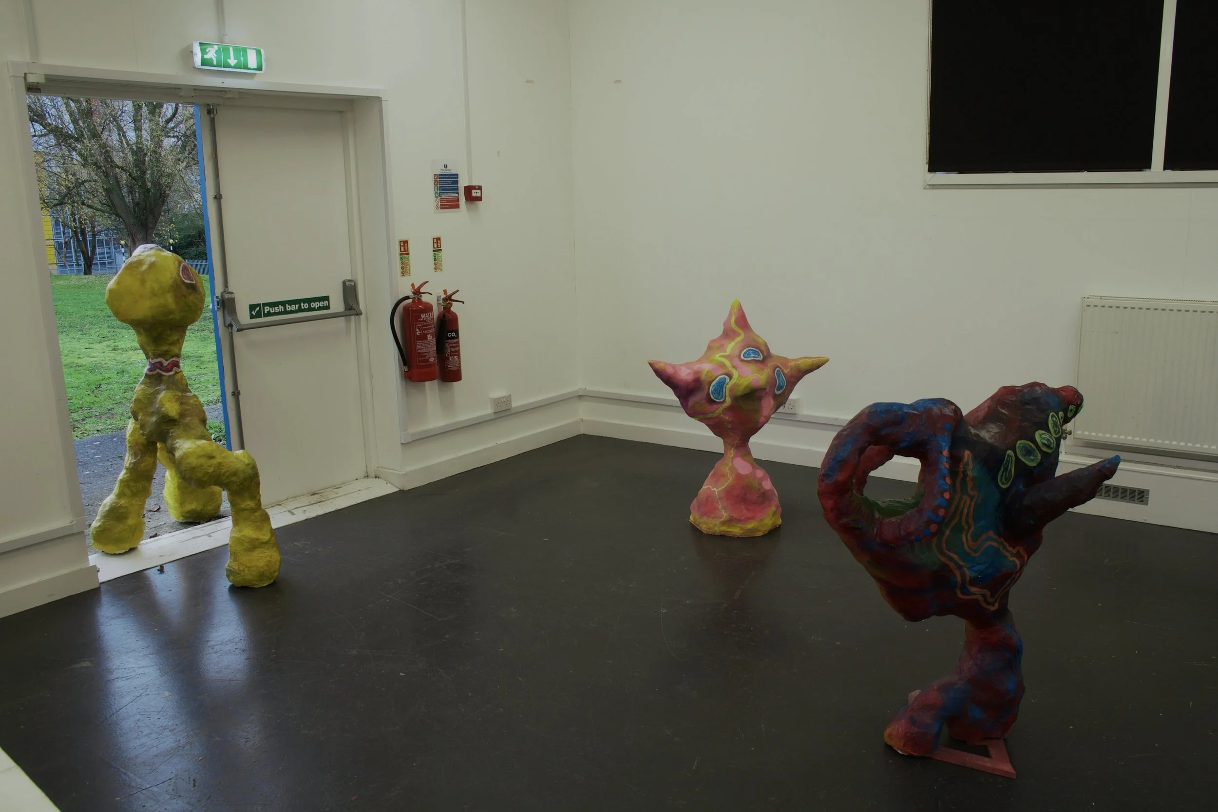 Three colorful abstract sculptures inside a room near a door leading outside to a grassy area with trees.