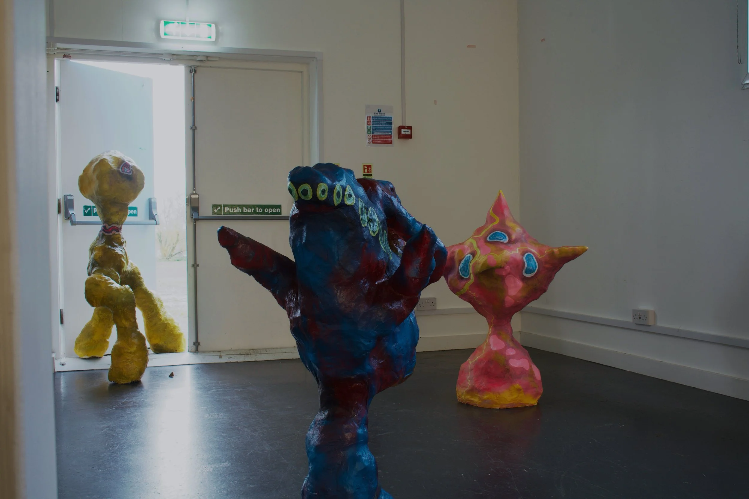 Three colorful abstract sculptures inside a room near a door with an exit sign.