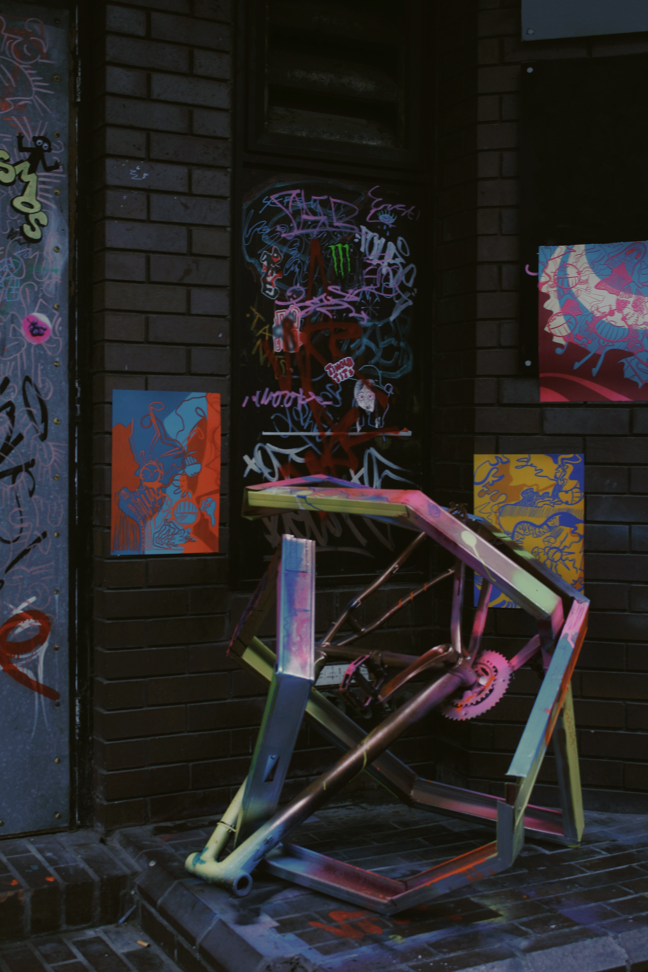 Street art display featuring colorful paintings and a metallic sculpture of a reclining person in front of a dark brick wall.