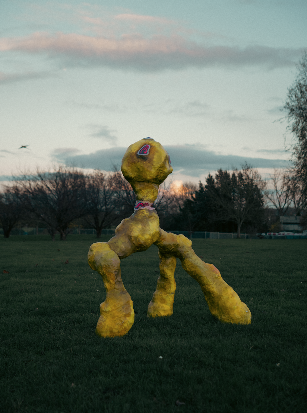 A yellow, abstract, sculpture-like figure with multiple limbs stands on a grassy field during sunset, with trees and a cloudy sky in the background.
