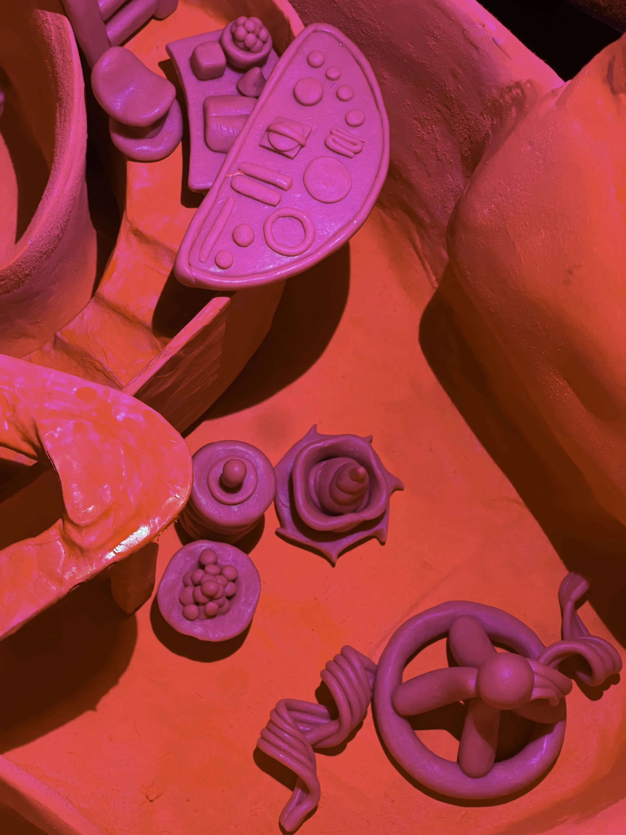 Close-up of pink clay toys, including a remote control, a slice of cake with round topping, a swirl of cream, a cluster of small spherical objects, and a mini steering wheel, on an orange surface under pink lighting.