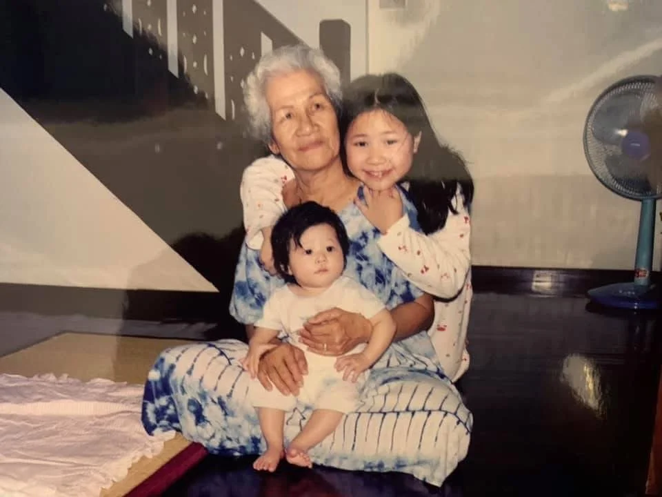 An elderly woman with white hair sitting on a cushion, holding two young children, one in each lap. One girl with long black hair and a smiling expression is sitting beside her. A boy with short black hair is sitting on her lap. They are inside a room with a dark hardwood floor, a standing fan, and a mural on the wall in the background.