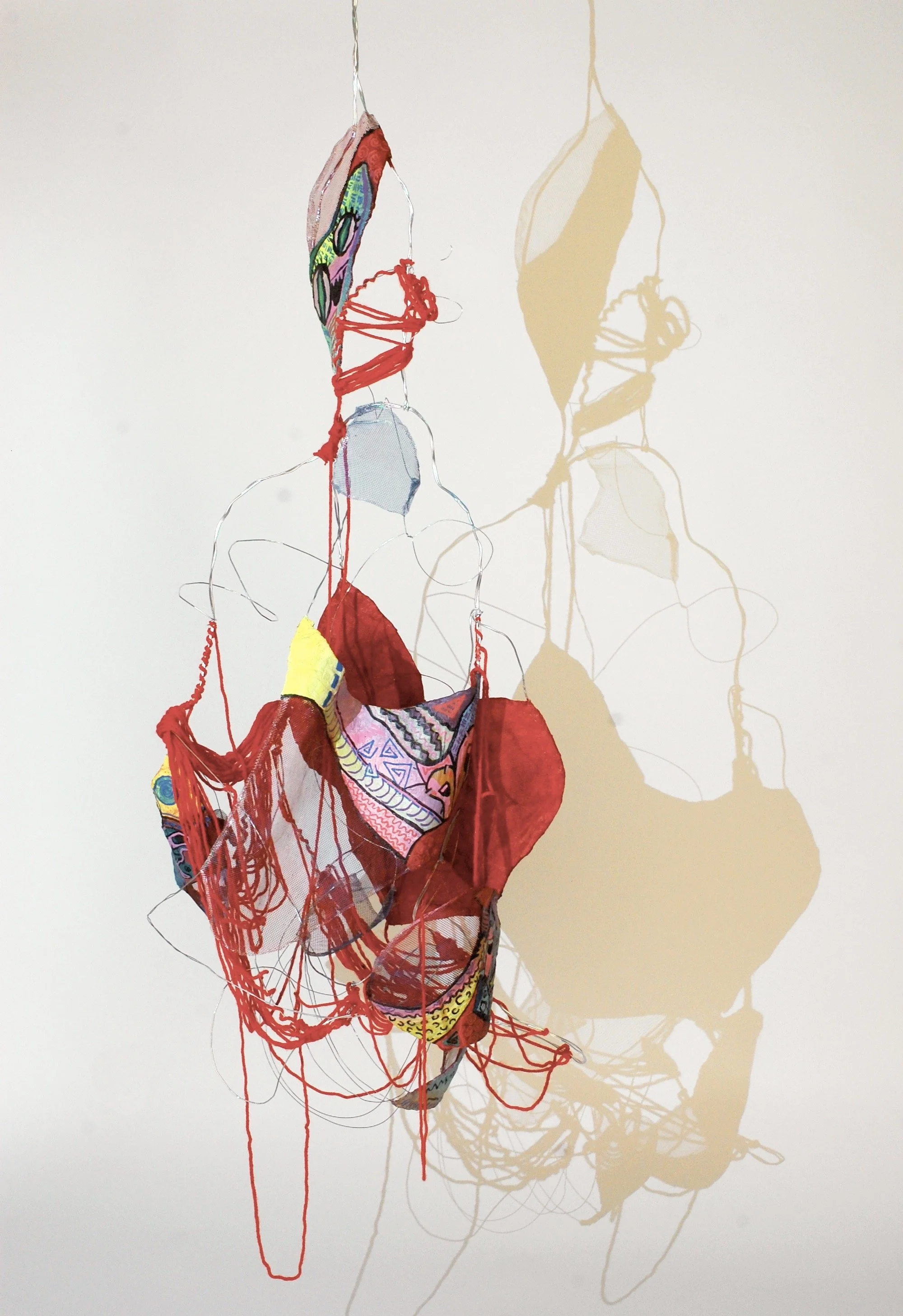 Colorful fabric and string sculptural art hanging against a plain wall, casting a shadow.