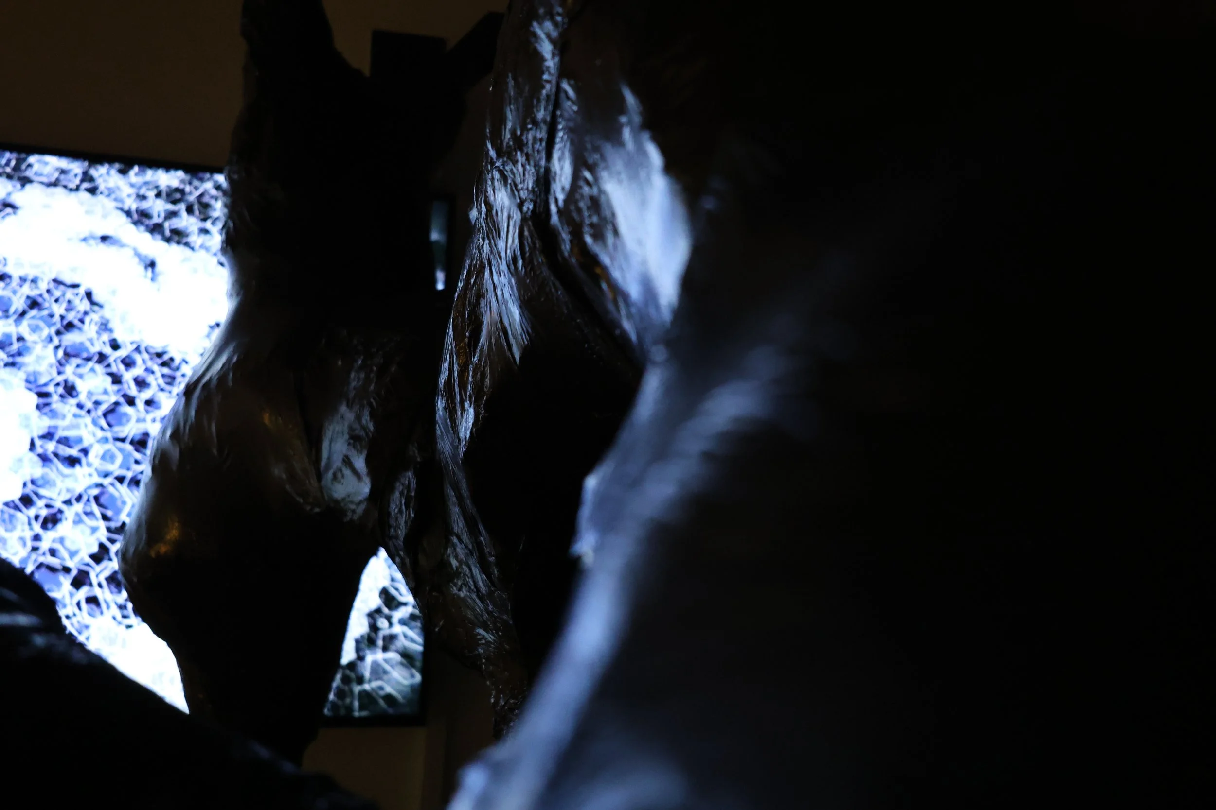 Close-up of dark, textured, reflective materials or sculptures in a dimly lit room, with a brightly illuminated digital screen in the background displaying a complex pattern.