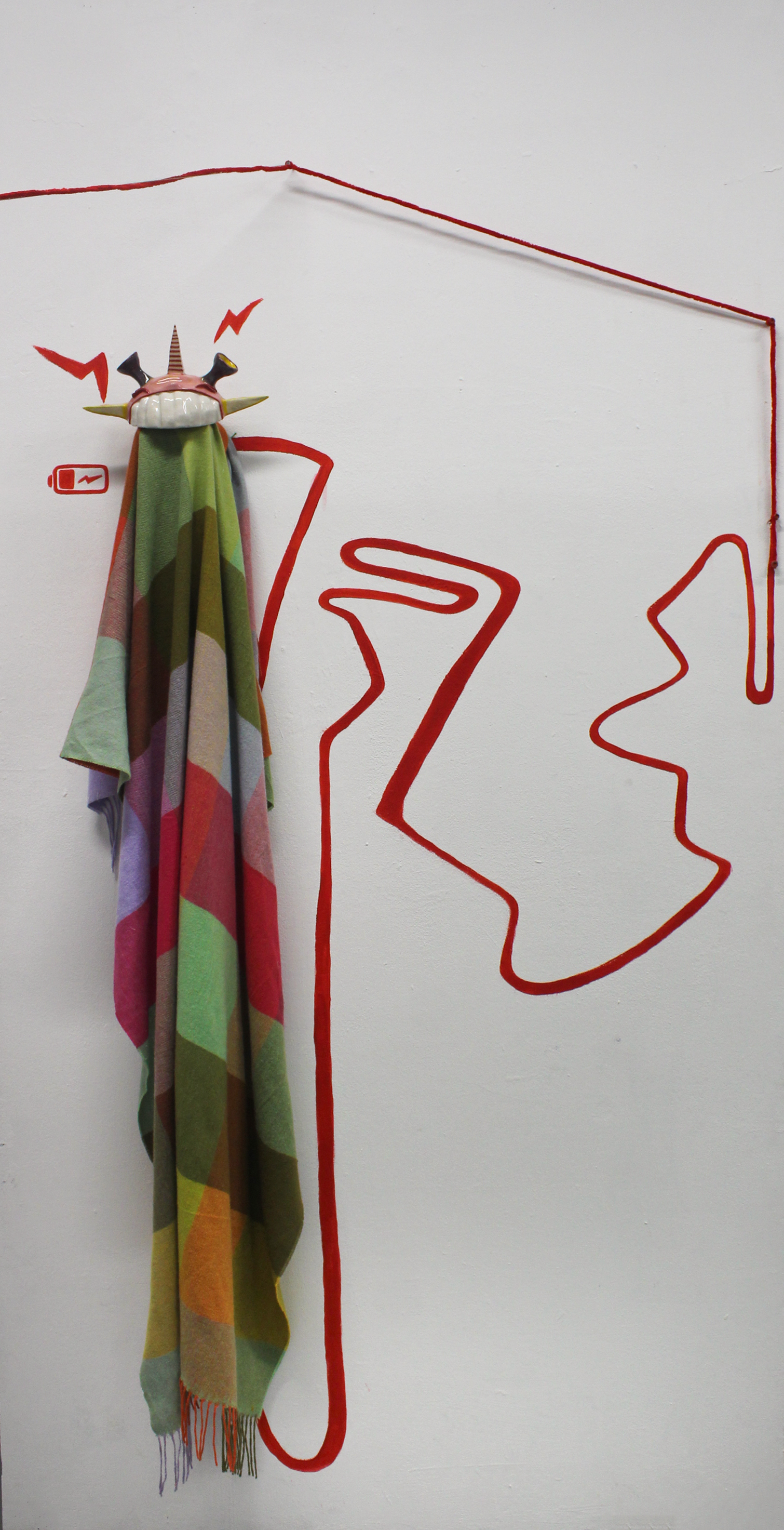 Wall art featuring a colorful fabric with an abstract face design in red outline, a small red battery icon, and a red squiggly line connected to the fabric.