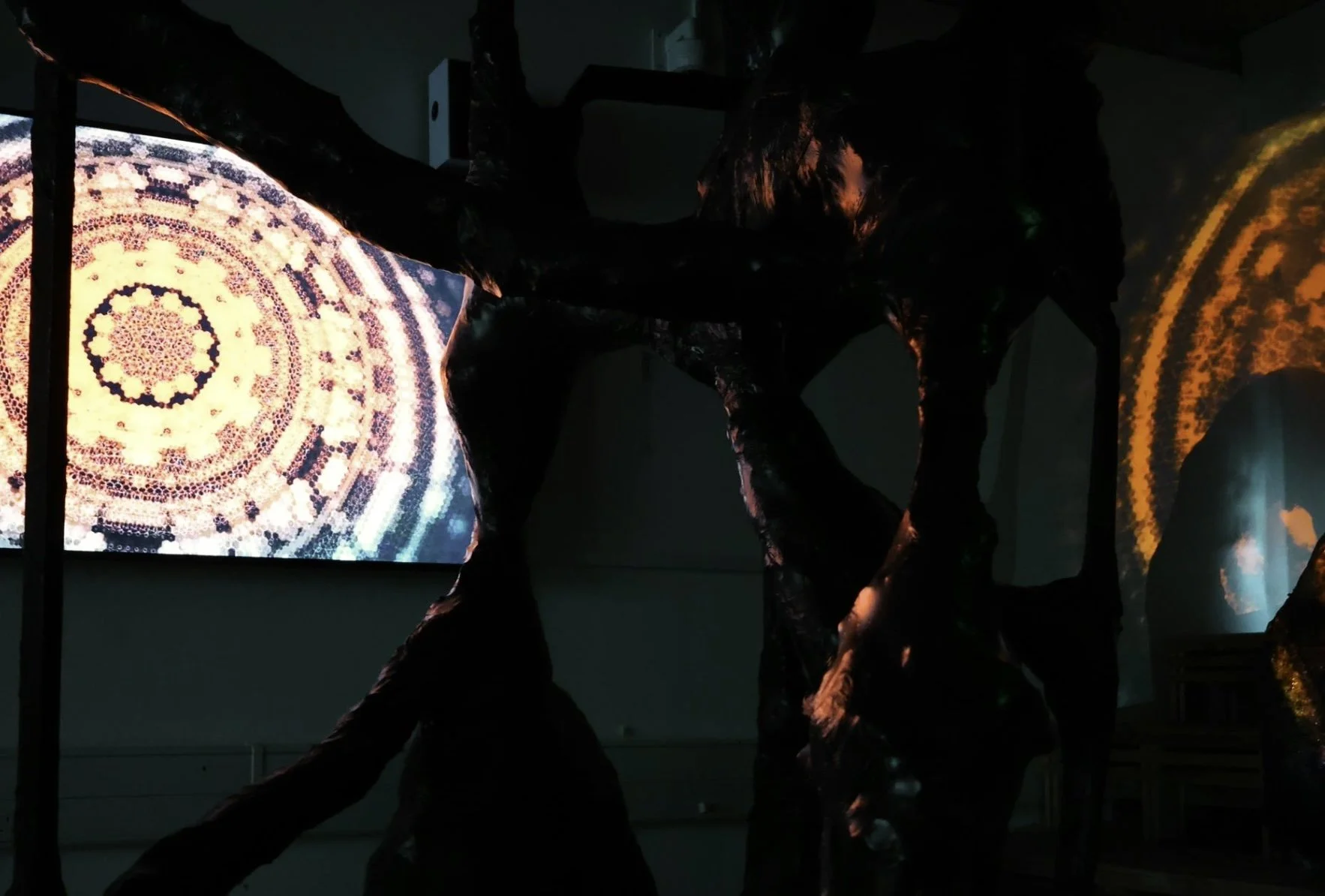 Dark art installation featuring a large abstract sculpture with intertwining limbs, with illuminated digital screens displaying intricate circular patterns in the background.