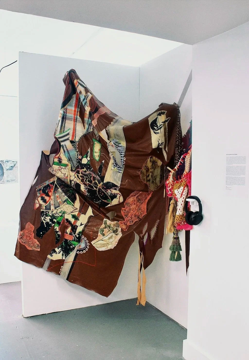 Abstract mixed media art installation with a fabric collage, headphones, and textile objects on a white wall in a gallery setting.