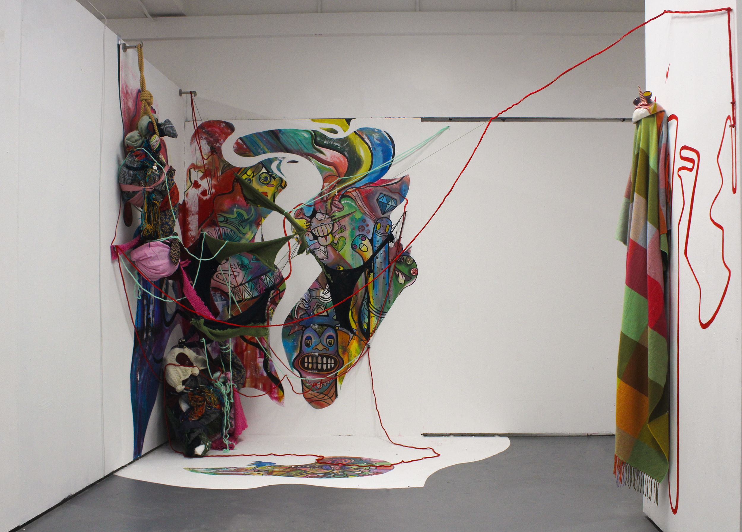 An art installation in a white room featuring a large, colorful, abstract mural with surreal and cartoon-like faces and figures. Various fabric and crocheted objects are attached to the walls, as well as red strings creating a web-like structure connecting elements. A colorful striped blanket is draped on the right wall, and a painted shape is on the floor beneath the mural.