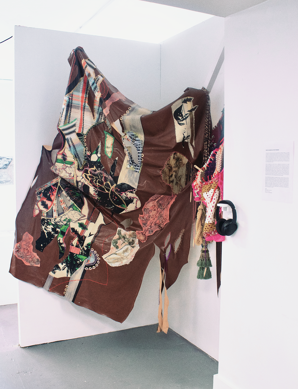 Mixed media art installation with a fabric piece comprising collage and textile elements, mounted on a white wall with headphones hanging nearby.