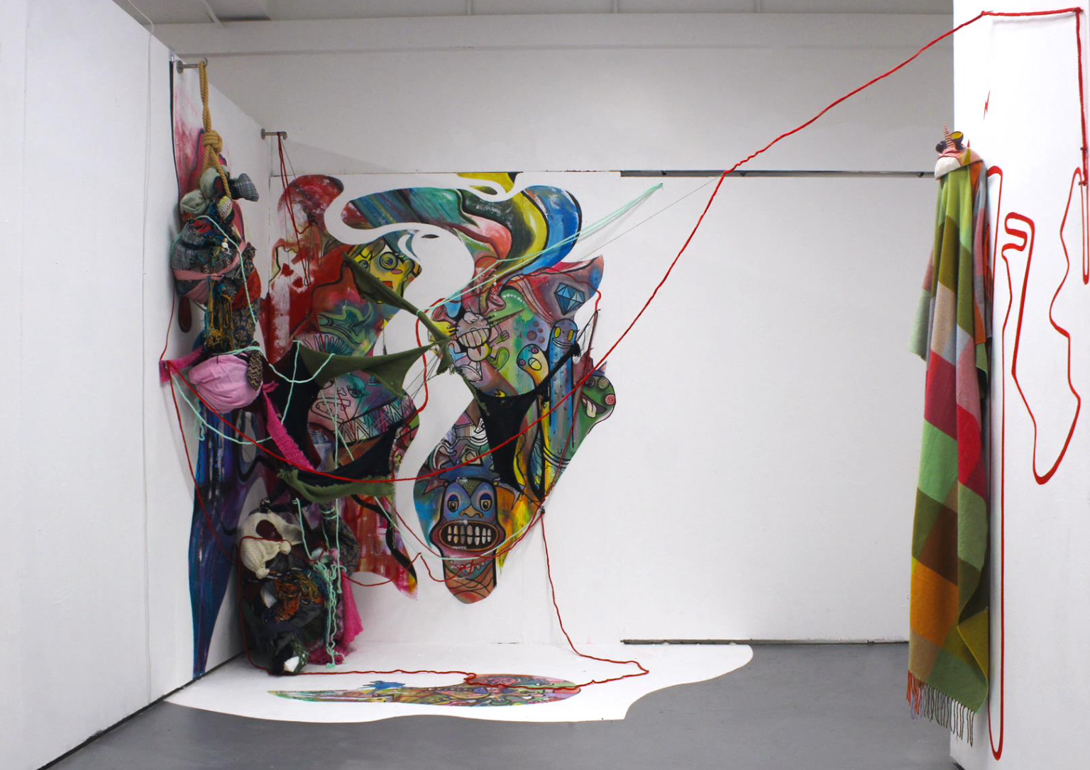 An art installation featuring a large, colorful abstract mask with various textures and elements like yarn, fabric, and painted surfaces, set against a white wall with hanging textiles and a wavy red thread.