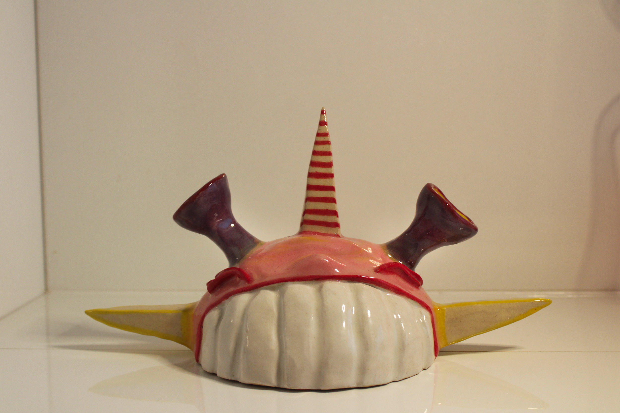 Colorful ceramic sculpture resembling a whimsical creature with a rounded body, two horn-like structures, a tall striped cone on top, and extended flat wings.