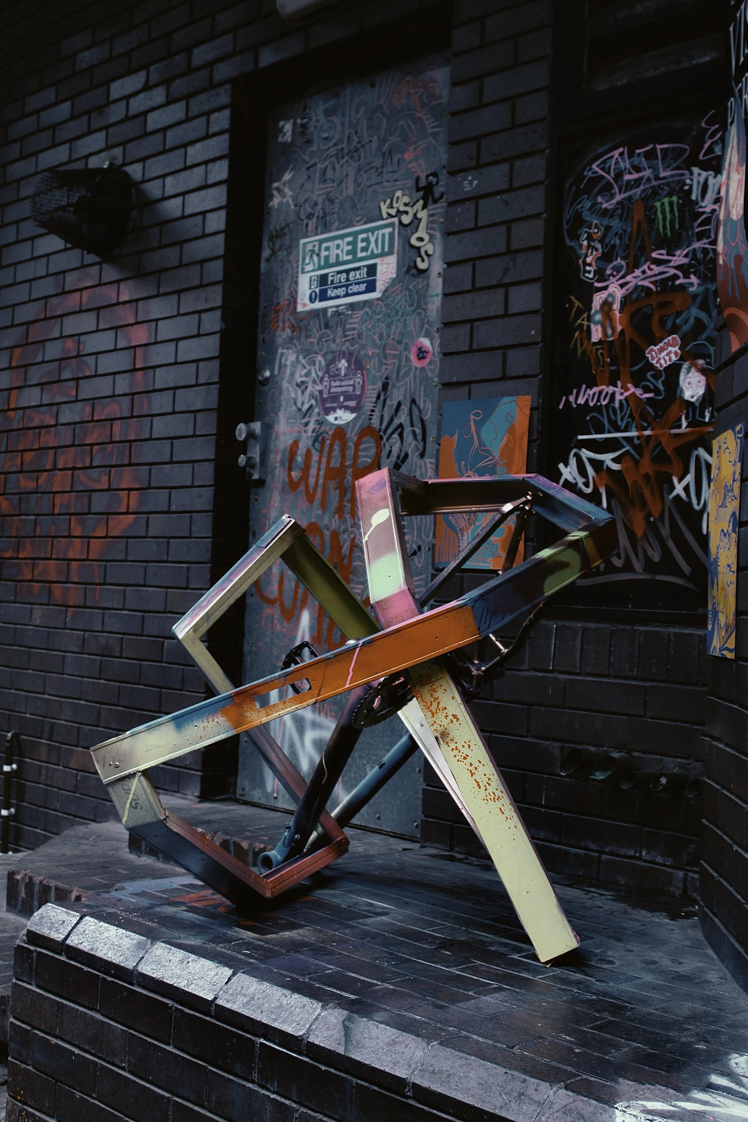 A broken, upside-down bicycle resting against a brick wall covered in colorful graffiti art. A fire exit door with stickers and graffiti is visible behind the bicycle.