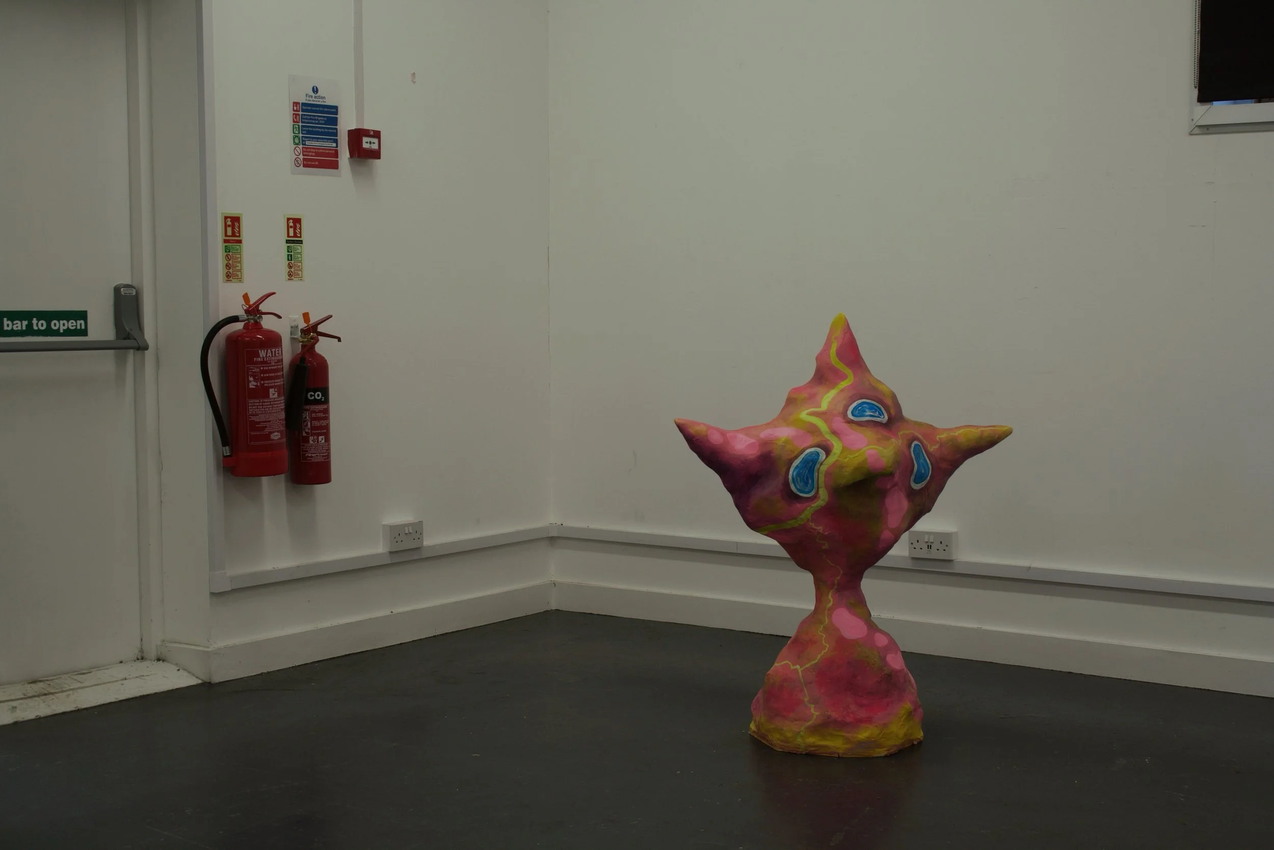 Abstract colorful sculpture in a gallery room with white walls, fire extinguishers on the wall, electrical outlets, and a window.