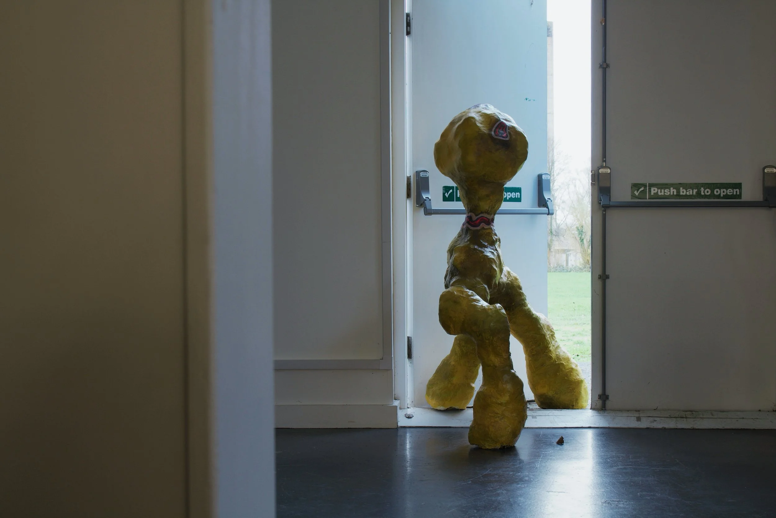 An abstract sculpture resembling a yellow, textured creature with a large head, standing at an open door.