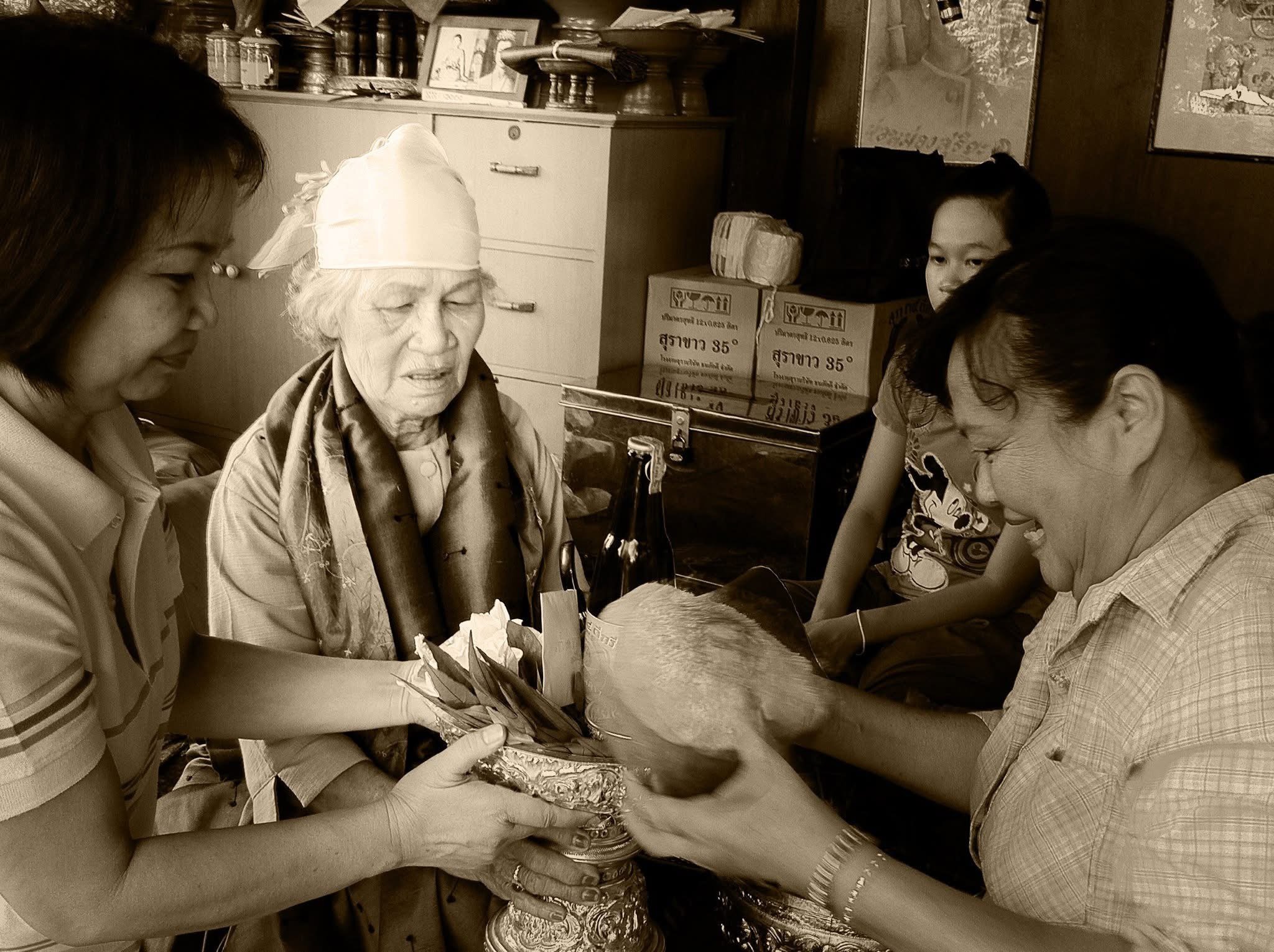 An elderly woman receiving a traditional offering from a group of women in a room decorated with framed pictures and boxes.