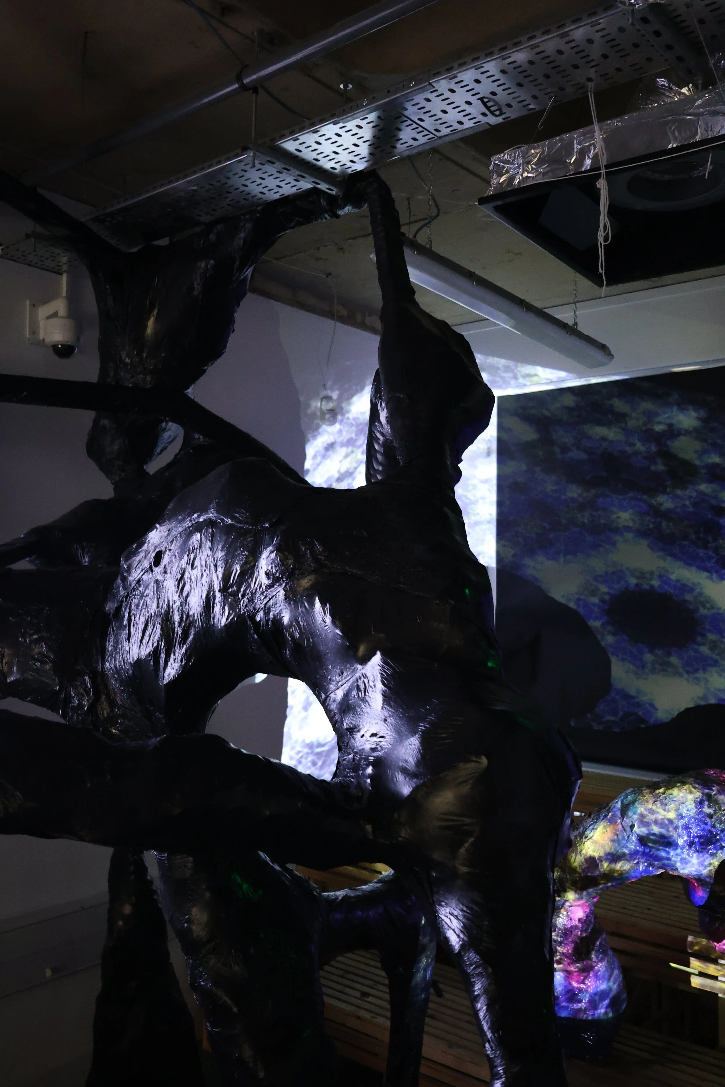 An abstract sculpture resembling a human figure reaching upward, illuminated with dark and colorful lights, in a dimly lit gallery or installation space.