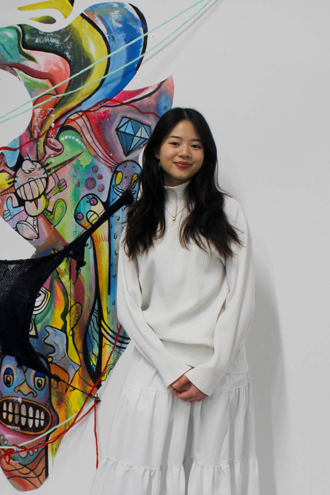 Young woman with long dark hair standing in front of a colorful graffiti mural on a white wall, wearing a white long-sleeve top and a white skirt, smiling slightly.