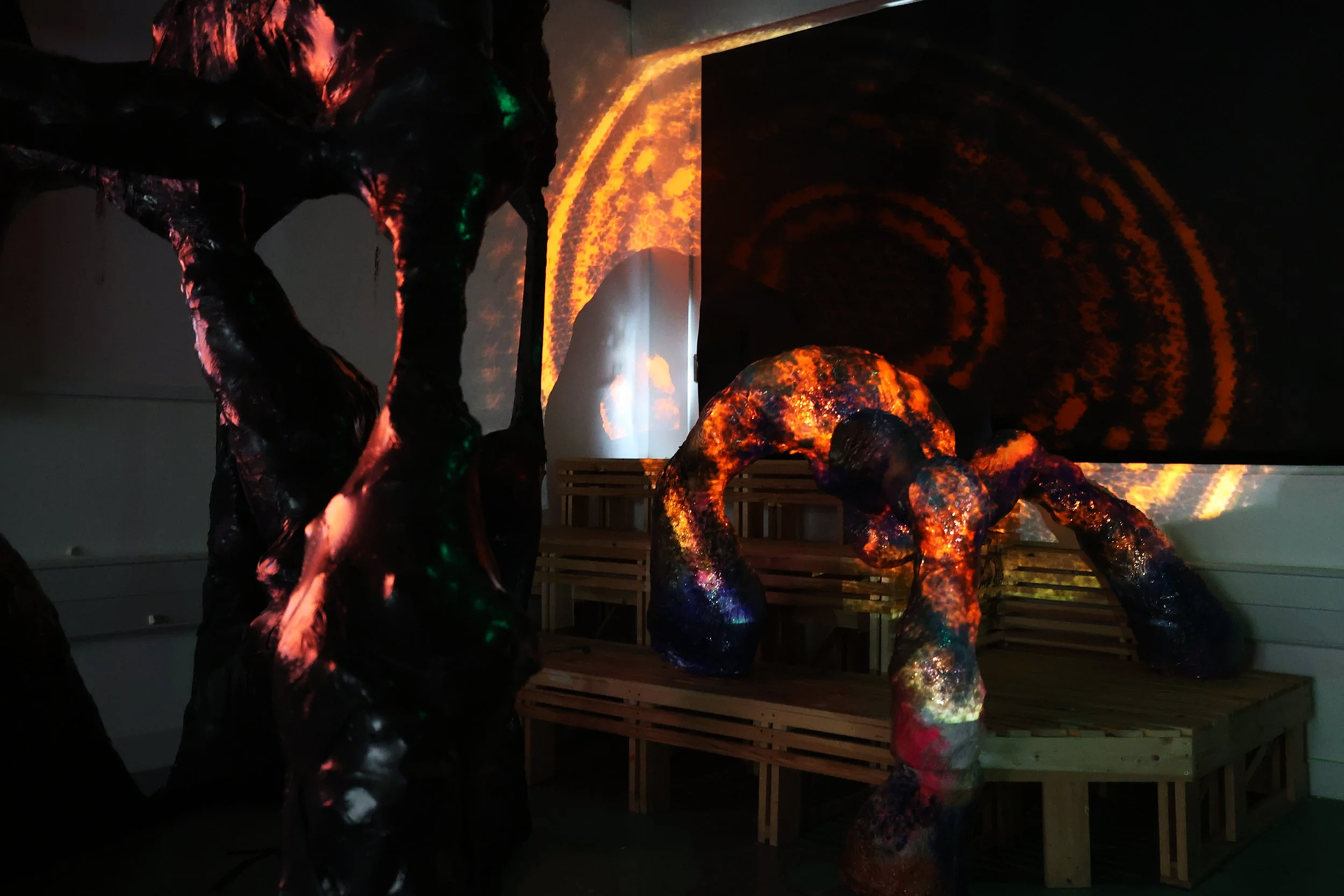 An art installation with abstract, shiny, black sculptures resembling organic shapes, illuminated by colorful lights, with a large swirling cosmic projection on the wall behind.