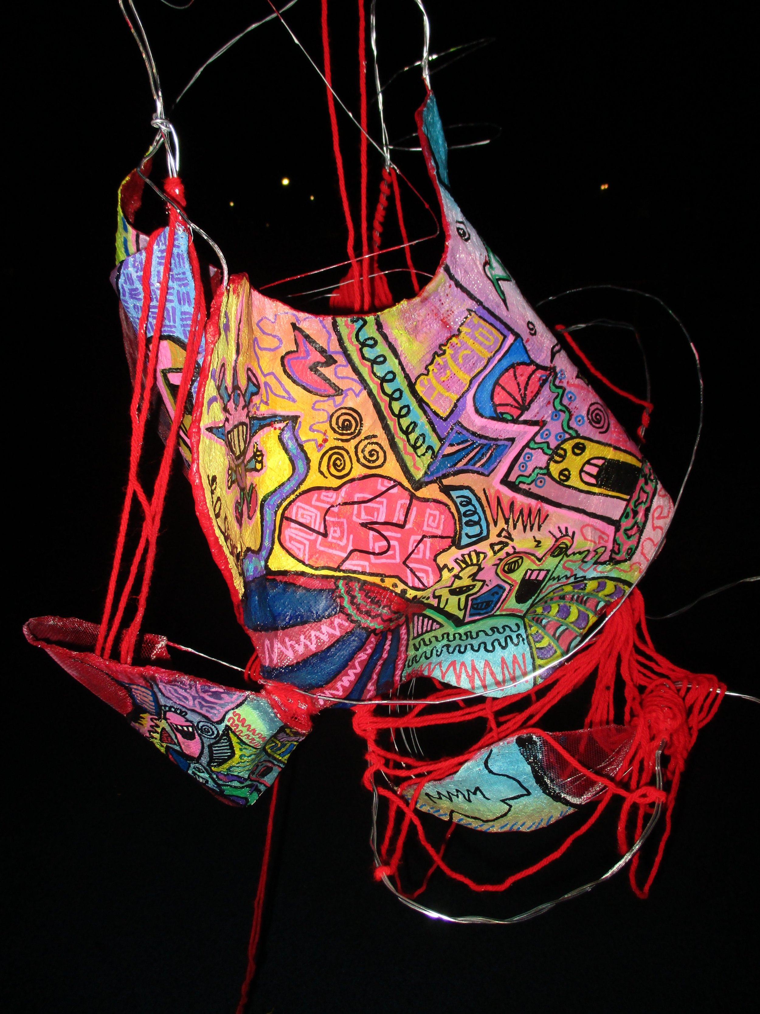 Colorful, abstract fabric mask with various doodles,文字, and shapes, adorned with red and silver strings and wires on a black background.