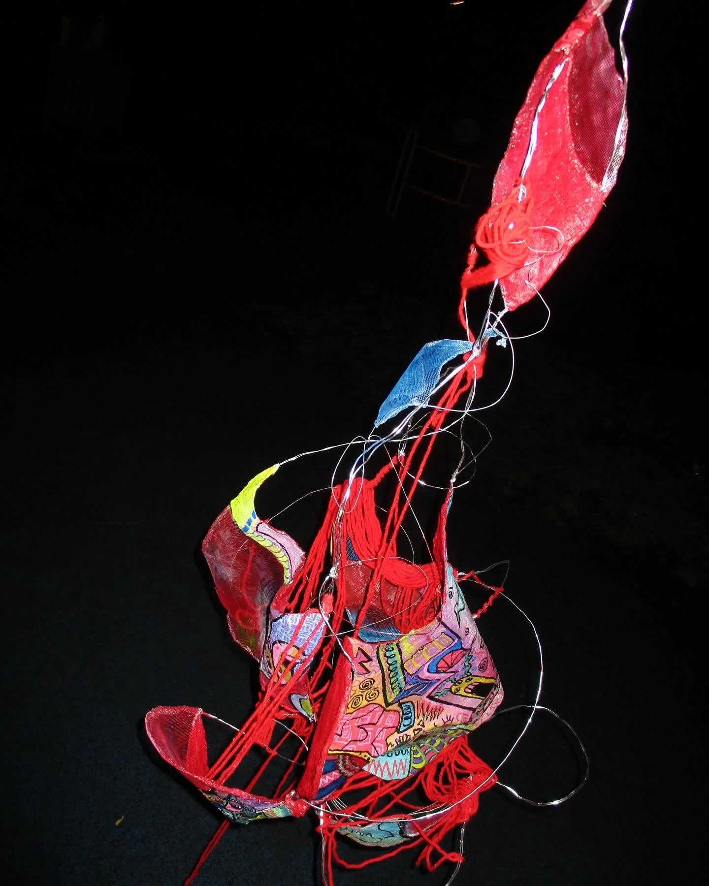 A tangled, colorful string art piece with red strings and pieces of fabric on a black background.